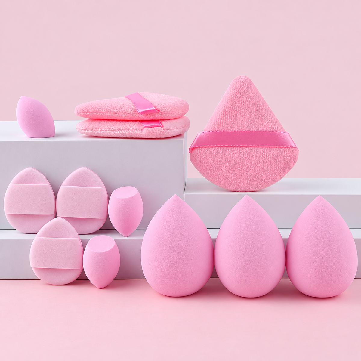 Rosa makeupsvampar och puffar uppställda på vita podier mot rosa bakgrund